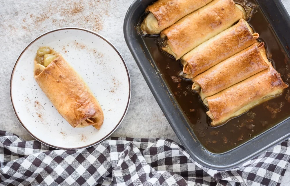 Freshly baked apple pie enchiladas on a white plate, dusted with cinnamon, with more in a baking tray alongside a checkered napkin.