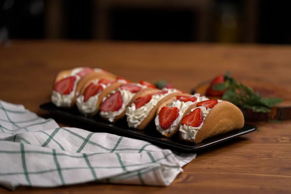 Plate of Strawberry Cheesecake Tacos, featuring golden-brown taco shells filled with creamy pink strawberry cheesecake filling, topped with fresh strawberry pieces, on a white plate over a neutral-toned cloth, offering a deliciously sweet twist on a classic treat.