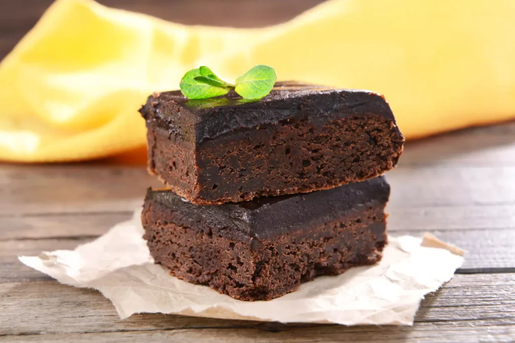 Two rich chocolate brownies with glossy frosting stacked on parchment paper, garnished with a fresh mint leaf, on a rustic wooden table with a yellow cloth in the background.