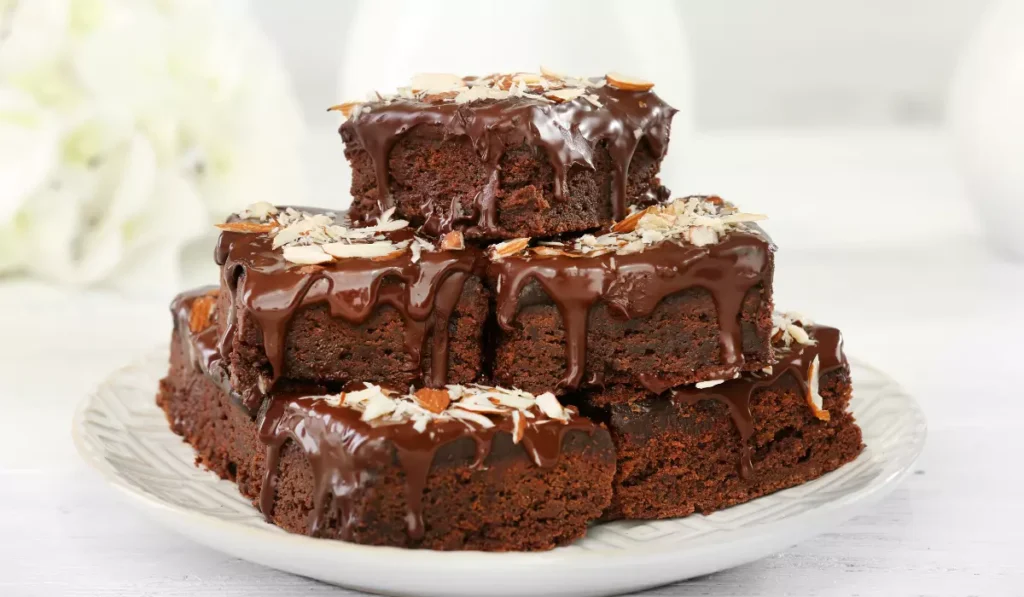 A stack of freshly baked brownies topped with a rich chocolate glaze and almond slivers on a white ceramic plate.