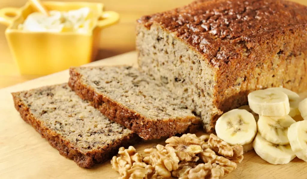 Sliced banana bread with walnuts and banana slices on a wooden cutting board, with a bowl of butter in the background.