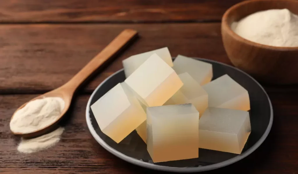 A bowl of translucent agar-agar cubes next to a wooden spoon of powder and a bowl of white powder on a rustic wooden table.