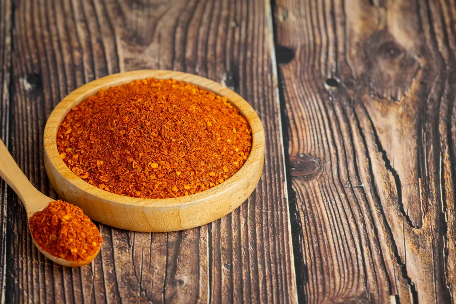A wooden bowl full of vibrant red chorizo seasoning alongside a spoonful of the spice blend on a rustic wooden table.