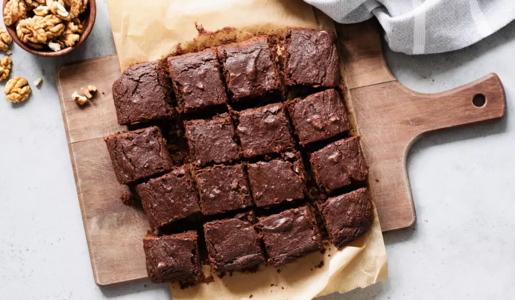 A wooden cutting board with a parchment-lined batch of freshly baked brownies cut into squares, accompanied by a bowl of walnuts on the side and a light grey cloth in the background.