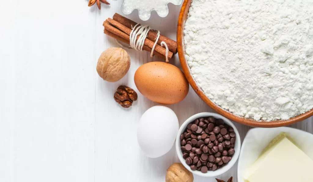 Assorted baking ingredients on a white surface, including flour, eggs, walnuts, cinnamon sticks, chocolate chips, and butter. Corn Starch Baking Powder