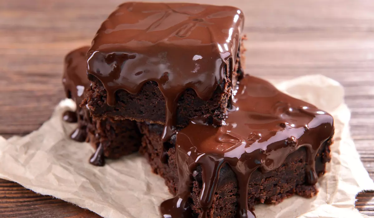 Two decadent chocolate brownies on parchment paper, drenched in glossy chocolate ganache, on a wooden surface.