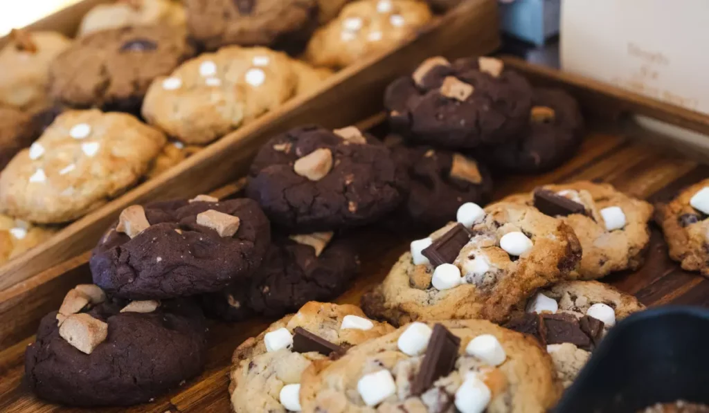 A variety of freshly baked cookies on display, including chocolate cookies with almond pieces and classic cookies with chocolate chunks and mini marshmallows, arranged neatly in a wooden tray.