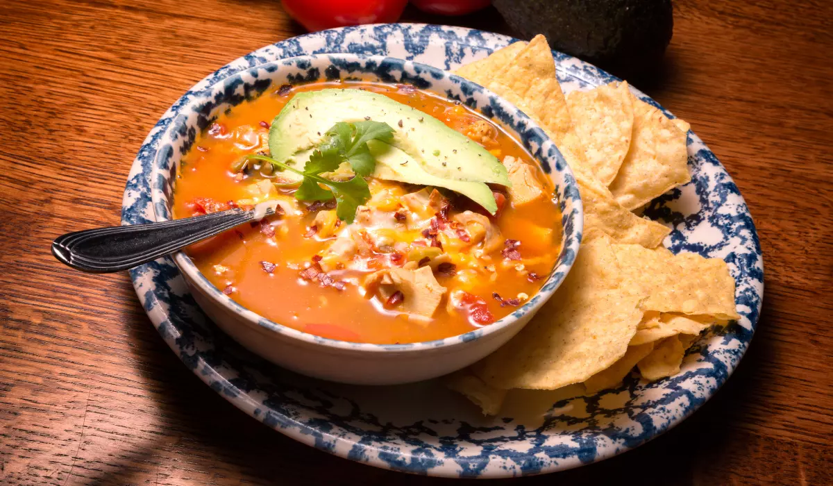 A bowl of chicken tortilla soup garnished with avocado slices and cheese, served with tortilla chips, on a patterned ceramic plate set on a wooden table.