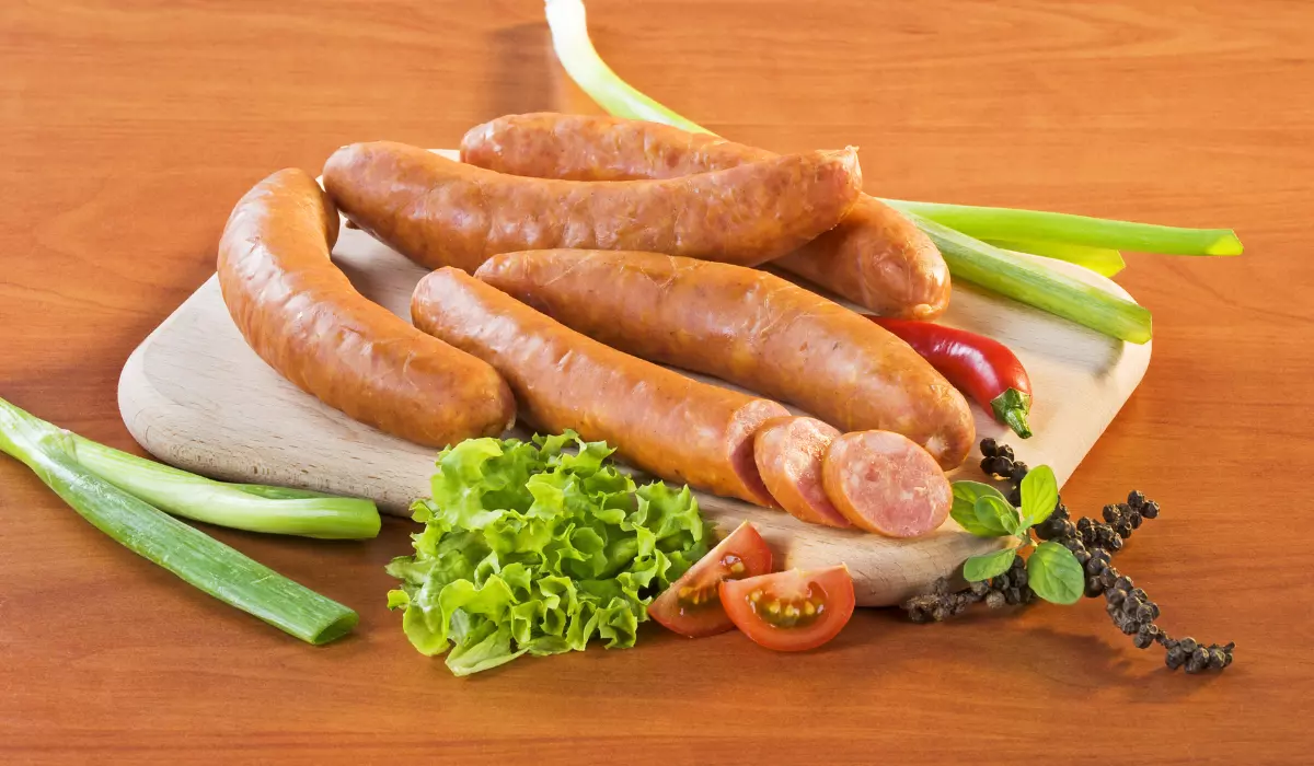 A wooden cutting board on a polished table holds a selection of kielbasa sausages with slices showing the juicy interior, accompanied by fresh green onions, lettuce, tomato, and a sprig of basil with a scatter of black peppercorns.