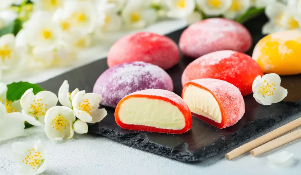 Assorted mochi ice cream on a slate board with jasmine flowers and chopsticks on a light textured surface.