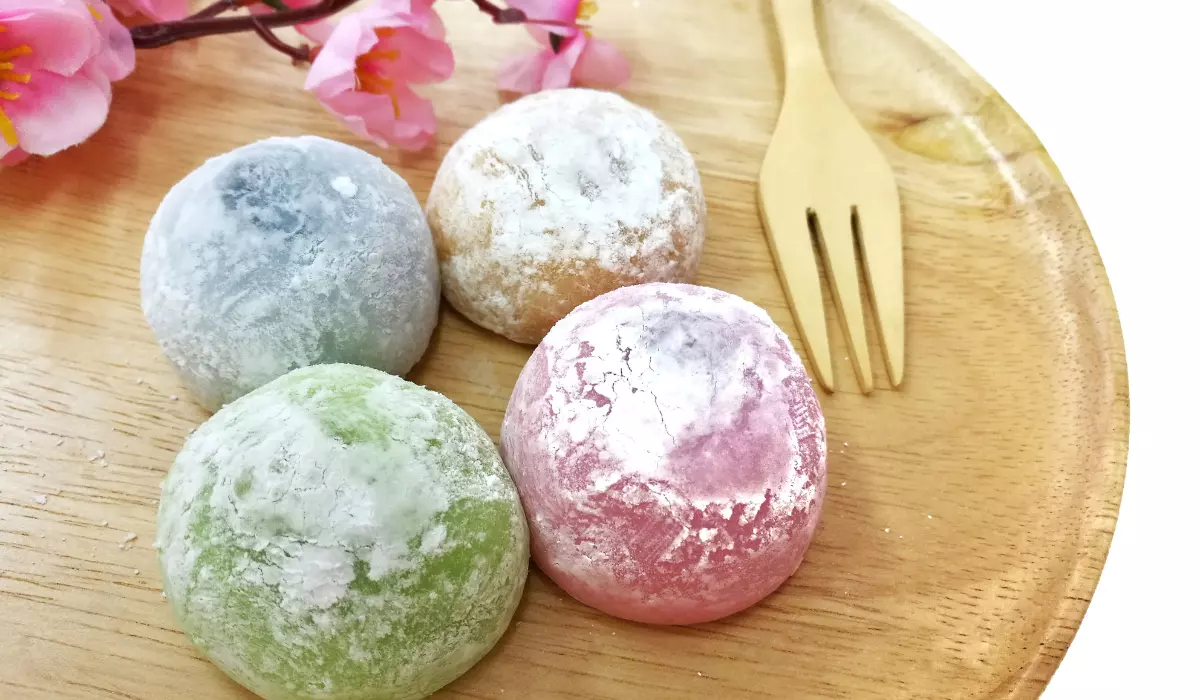A wooden plate with four colorful mochi dusted with flour alongside a wooden fork and artificial pink flowers.