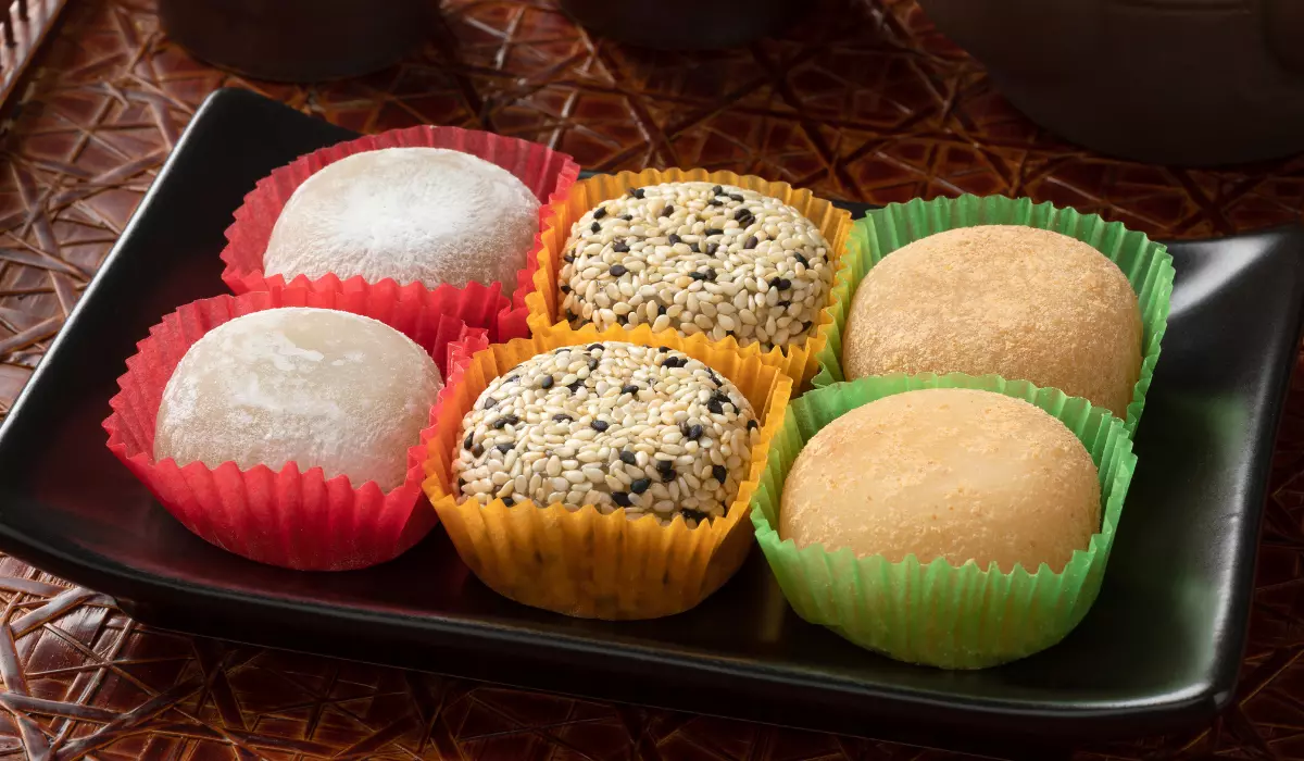 Four pieces of potato mochi in colorful cupcake liners on a black plate with a woven texture background.