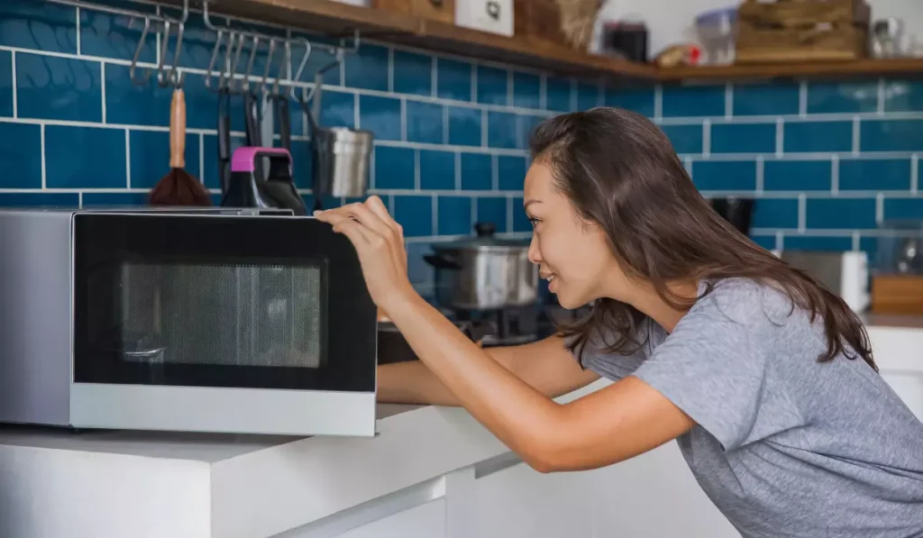 A woman excitedly looks into a microwave as she reheats a plate of brownies in a cozy home kitchen with blue tiles.