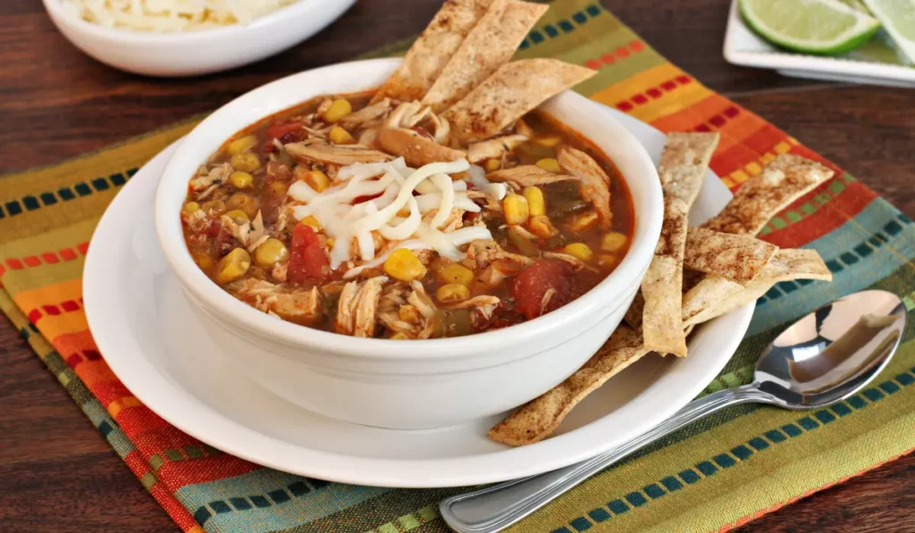 A bowl of chicken tortilla soup topped with shredded cheese, with a side of baked tortilla strips on a colorful striped placemat.