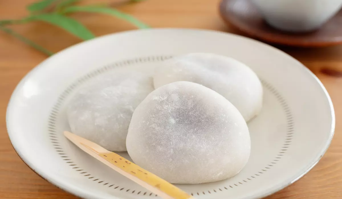 Three freshly made potato mochi on a ceramic plate with a wooden pick on the side, set on a wooden table with soft-focus greenery in the background.